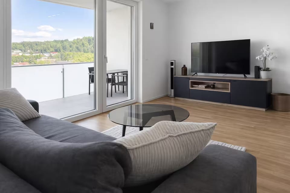 Helles Wohnzimmer mit Sofa, Glascouchtisch und TV-Möbel, dahinter die Balkontür mit Sitzplatz und Blick ins Grüne
