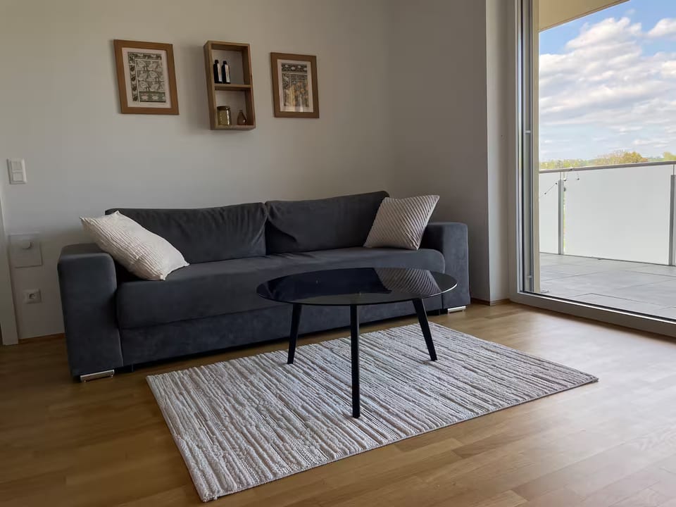 Graues Sofa mit Kissen und rundem Couchtisch auf hellem Teppich, rechts die Glastür zum Balkon
